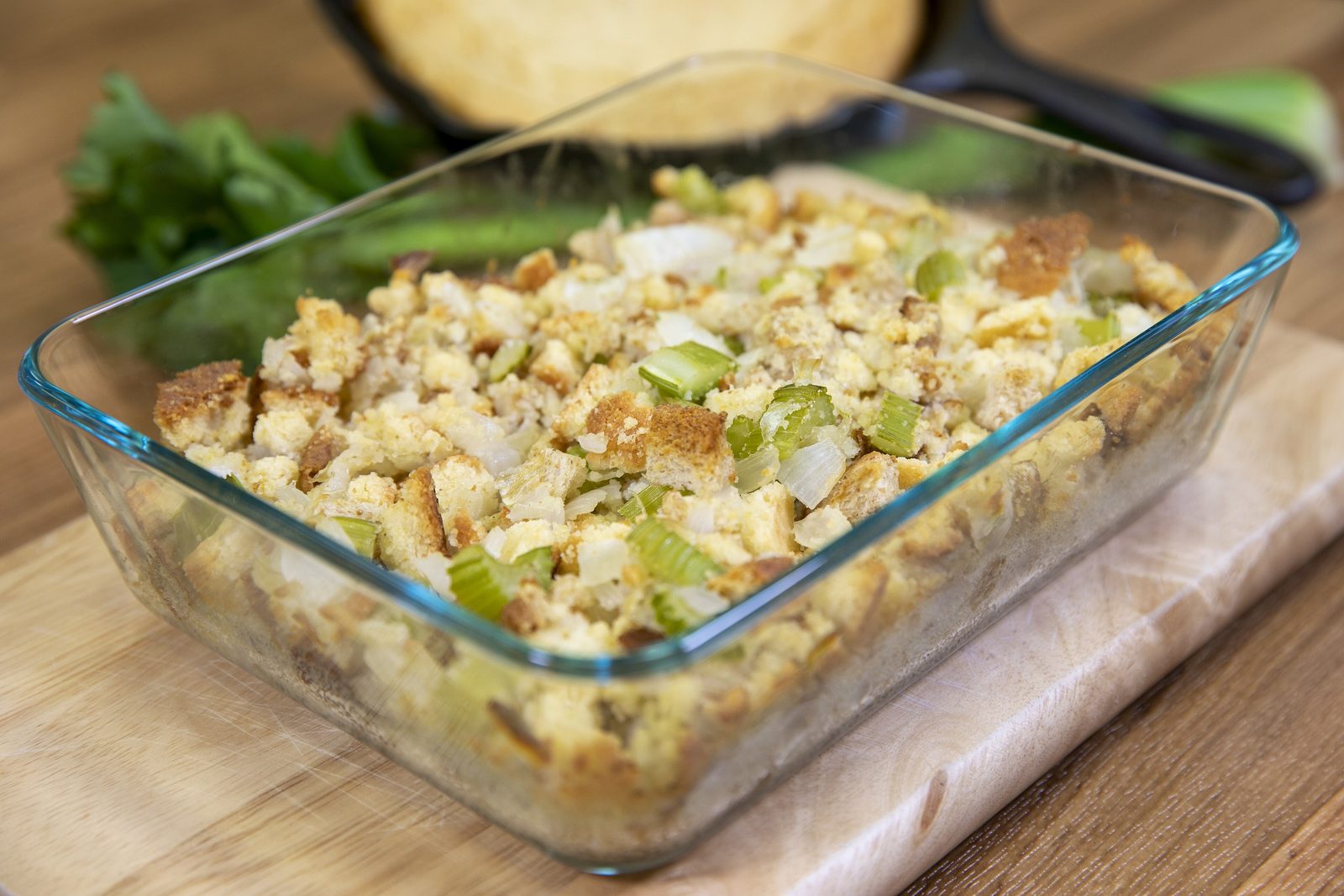 Baked cornbread dressing with celery and onion in a glass baking dish on a wooden board, a skillet of cornbread behind it