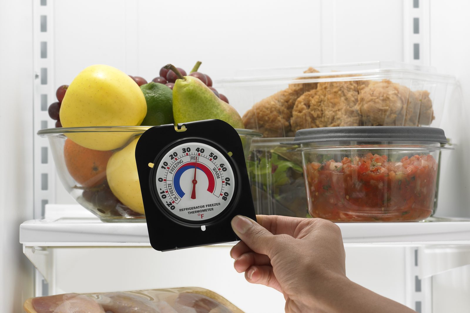 A hand holding a dial refrigerator thermometer in front of a fridge shelf with fruit, fried chicken and salsa
