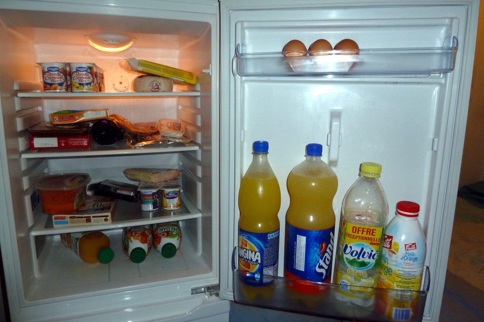 An open refrigerator with yogurt, cheese and leftovers on the shelves, eggs and four large juice bottles in the door