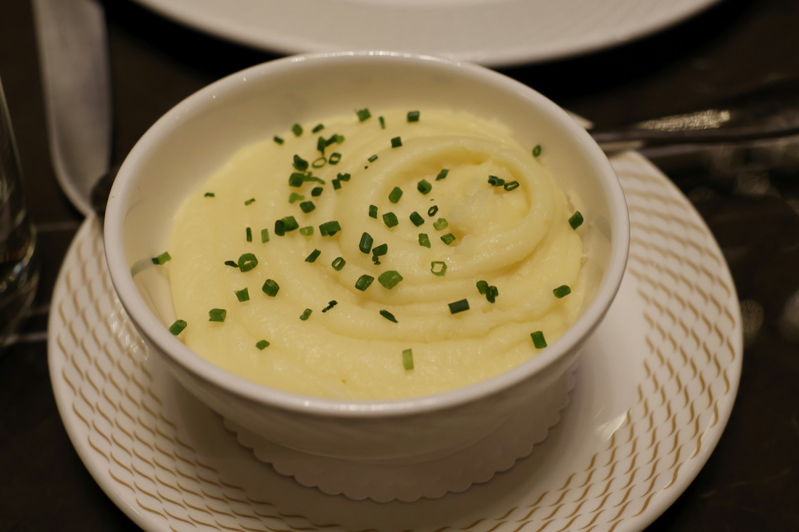 A white bowl of smooth mashed potatoes swirled and topped with chopped chives