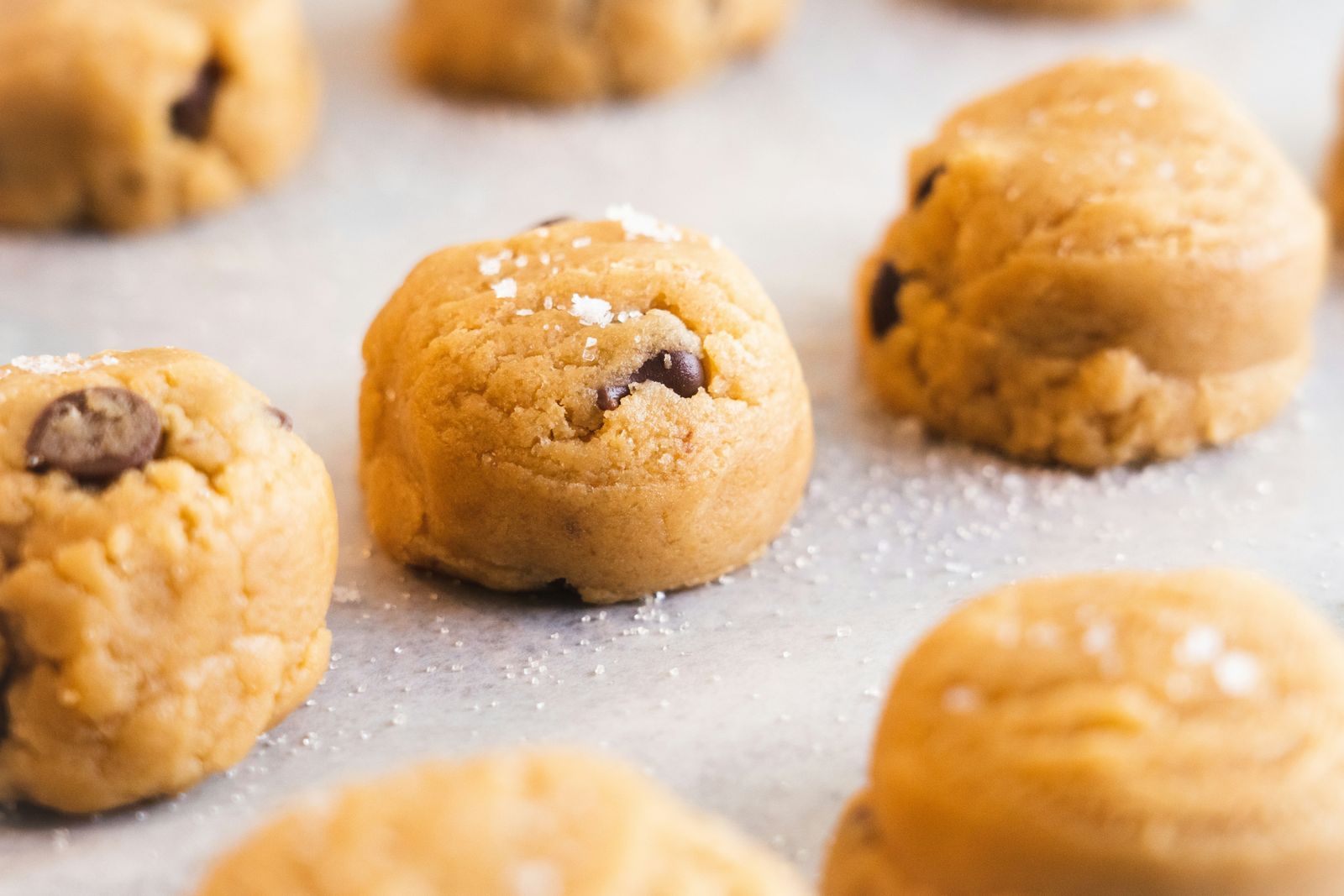 Balls of raw chocolate chip cookie dough sprinkled with sea salt on a parchment-lined baking sheet, waiting to go in the oven
