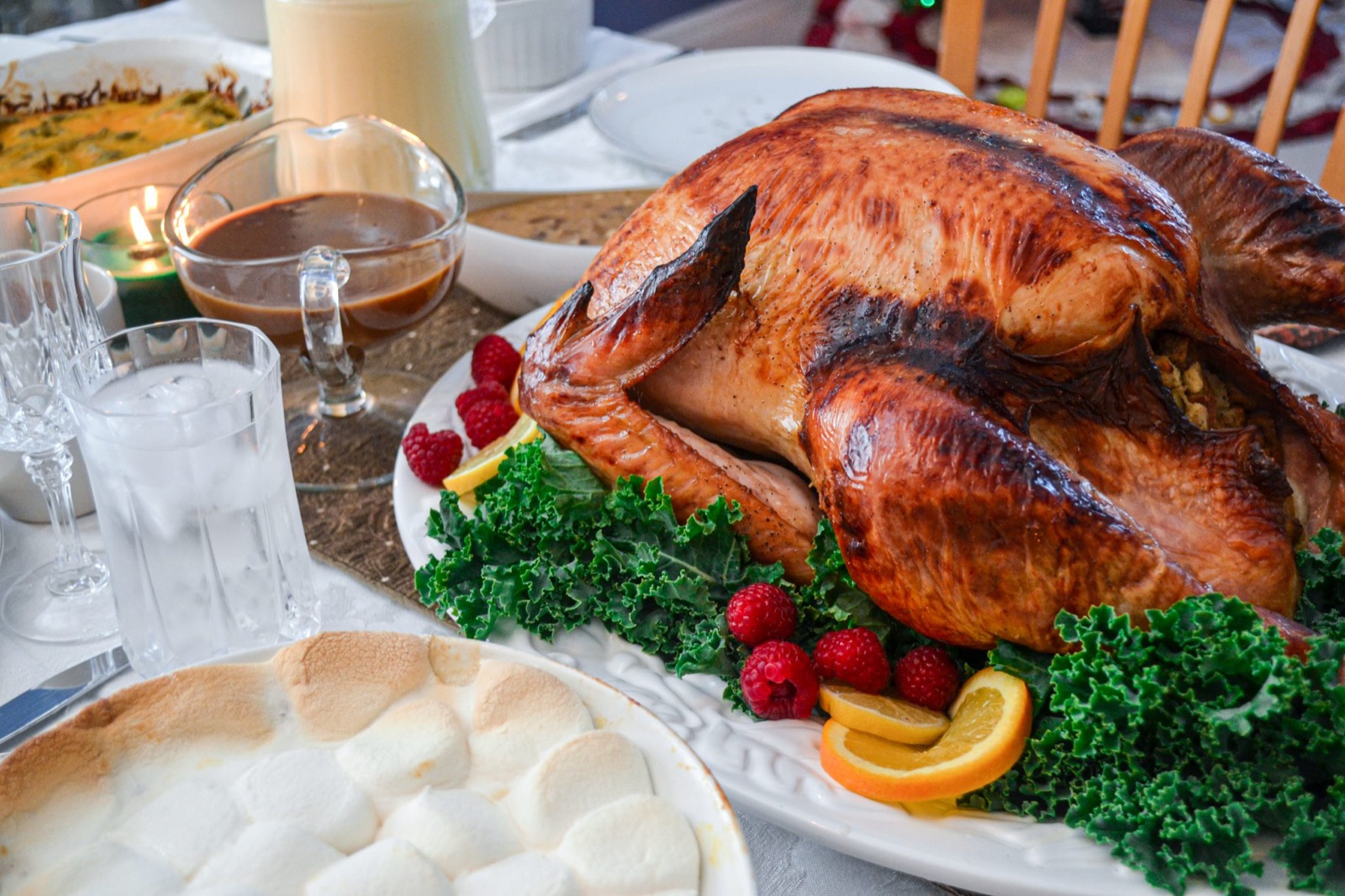 A roast stuffed turkey on a platter with kale, raspberries and orange slices, a boat of gravy, a green bean casserole and sweet potatoes topped with toasted marshmallows